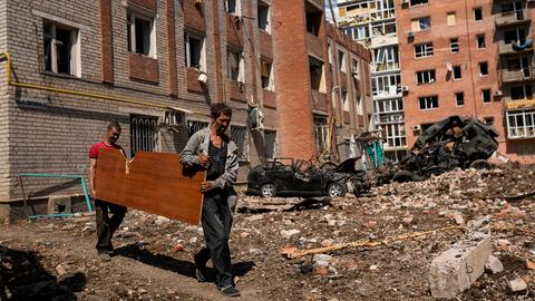 Zwei Männer tragen eine Holzplatte neben schwer beschädigten Gebäuden und zerstörten Autos in Bachmut (Ukraine). (dpa)