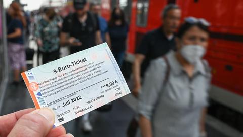Ein 9-Euro-Ticket für Juli 2022 wird am Hauptbahnhof hochgehalten, während Zugreisende aus einer Regionalbahn aussteigen ()