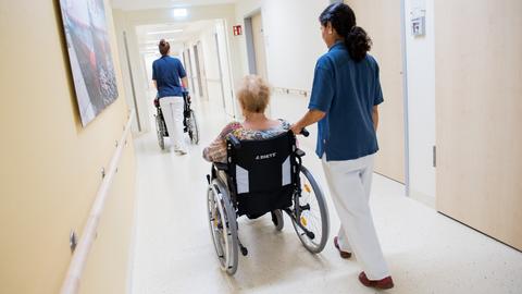 Zwei Pflegerinnen schieben Patienten in Rollstühlen über den Flur eines Krankenhauses.