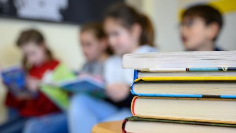 Auf einem Tisch liegen mehrere Bücher aufeinander gestapelt, im Hintergrund sieht man verschwommen Kinder beim Lesen.