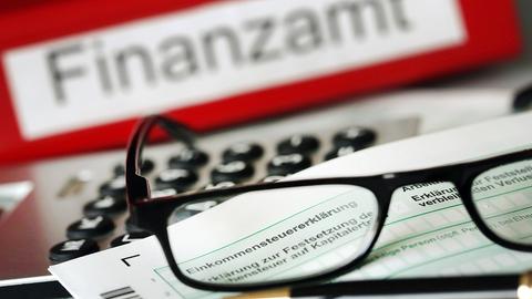 Auf einem Vordruck für die Steuererklärung liegt vor dem Aktenordner mit dem Aufdruck "Finanzamt" ein Stift und eine Brille.