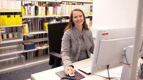 Frau sitzt an einem Computer, im Hintergrund die Regale eines Archivs.
