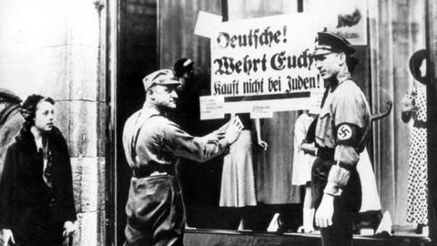  SA-Männer kleben ein Plakat mit der Aufschrift "Deutsche! Wehrt Euch! Kauft nicht bei Juden" an die Schaufensterscheibe eines Geschäfts, das in jüdischem Besitz ist 