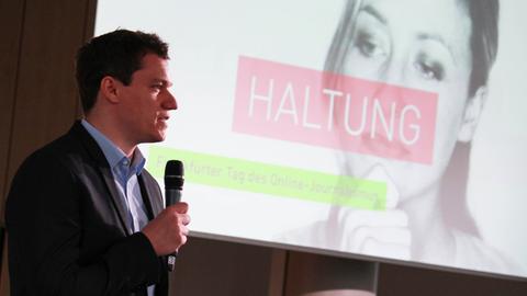 Moderator mit Mikrofon in der Hand vor einer Leinwand mit dem Schriftzug "Haltung"