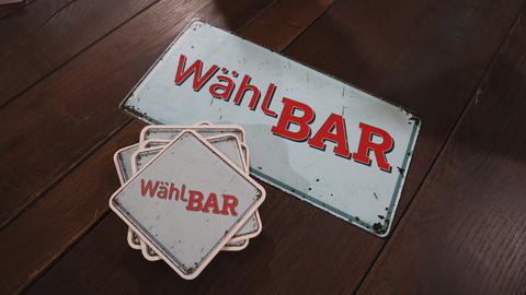 Bildmarke zu „WählBAR – Theken-Talk zur Bundestagswahl“. Ein Metallschild und Bierdeckel, bedruckt mit dem Begriff Wählbar, liegen auf einem Holztisch.