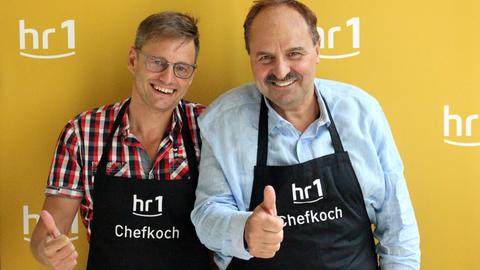 Spitzenkoch Johann Lafer (r) und Moderator Uwe Berndt (l) tragen Schürzen mit der Aufschrift "hr1-Chefkoch" und lächeln in die Kamera