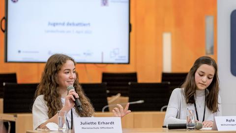 Jugend debattiert – Landesfinale Hessen 2022: Zwei junge Schülerinnen bei der Debatte - eine spricht ins Microphone und die Andere macht sich Notizen.