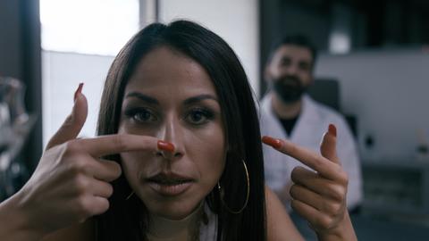 Negah Amiri zeigt auf ihre Nase, im Hintergrund sieht man verschwommen einen Mann im Arztkittel 