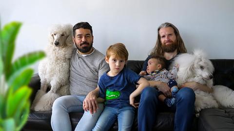 Das Paar Francisco (li.) und Chris (re.) sitzen mit ihren Kindern und Hunden auf einer Couch.