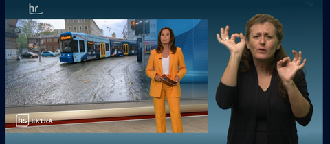 Eine Szene aus dem "hessenschau extra" zum Thema Starkregen. Moderatorin Claudia Schick im Studio, im Hintergrund ein Buld von einer überschwemmten SDtraße in Kassel. Im rechten Bildteil sieht man eine Gebärdendolmetscherin.