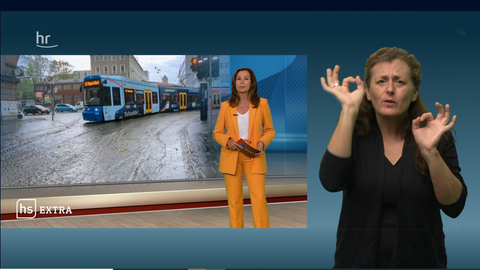Eine Szene aus dem "hessenschau extra" zum Thema Starkregen. Moderatorin Claudia Schick im Studio, im Hintergrund ein Buld von einer überschwemmten SDtraße in Kassel. Im rechten Bildteil sieht man eine Gebärdendolmetscherin.