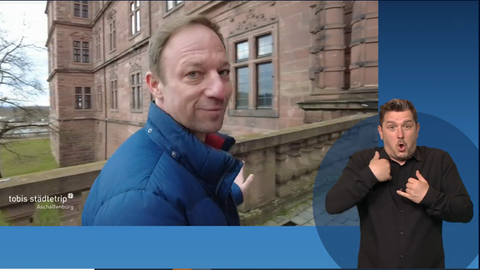 Tobias Kämmerer geht über eine Brücke auf ein historisches Schloss zu, von dem ein Gebäudeteil im Hintergrund zu sehen ist. Im rechten Bildteil ist ein Gebärdendolmetscher zu sehen.