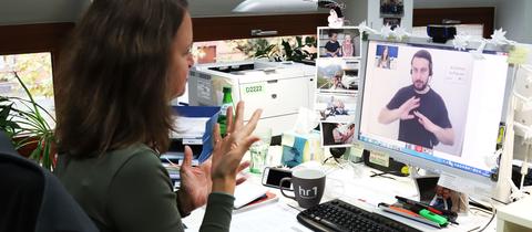Eine gehörlose Frau gebärdet vor einem PC-Monitor - sie telefoniert. Auf dem Monitor ist ein Gebärdensprachdolmetscher zu sehen sowie die hörende Anruferin.