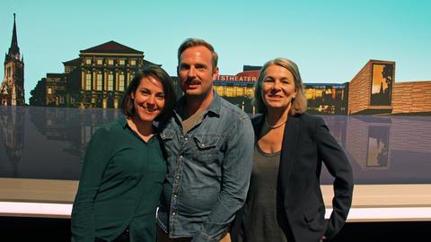 Die Komparsen Pauline Meignan, Martin Schlemmer und Ethel Dadam stehen vor der Kulisse im Fernsehstudio.