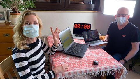 Moderatorin Franziska Reichenbacher und Produktionstechniker Uwe Tapken sitzen mit Mund-Nasen-Masken auf beiden Seiten eines kleinen Tisches mit drei geöffneten Laptops (zu sehen sind die Lotto-Grafik, das Kamerabild und ein Timecode) im Licht eines Scheinwerfers von hinten rechts.