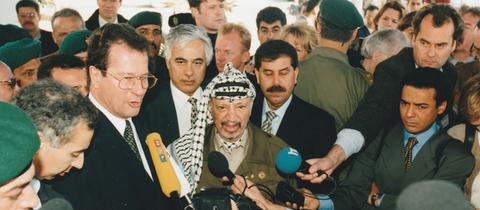 Der frühere Außenminister Klaus kinkel und Palästinenserführer Jassir Arafat umringt von einem Pulk Journalist*innen mit mikrofonen, darunter auch Christopher Plasss mit hr-Mikrofon, Aufnahme von 1997