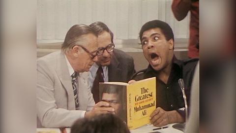 Muhammad Ali umringt von Reportern hält ein Buch in der Hand und zieht eine Grimasse