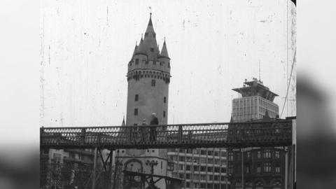 Schwarz-Weiß-Aufnahme des Eschenheimer Turms. Im unteren Drittel ist eine stählerne Fußgängerbrücke vor dem Turm zu sehen, auf der Menschen laufen.