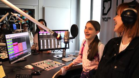 im YOU FM-Studio: Ein Mädchen am Moderationspult steht vor einem Sprechermikrofon und lacht. Neben ihr eine Moderatorin mit Kopfhörern, auch sie lacht.
